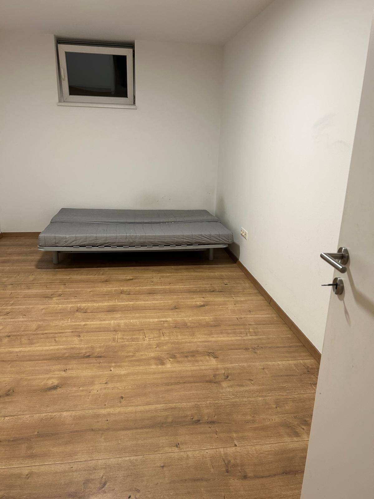 Einfaches Schlafzimmer mit Laminatboden, einem Bett und einem kleinen Fenster.
