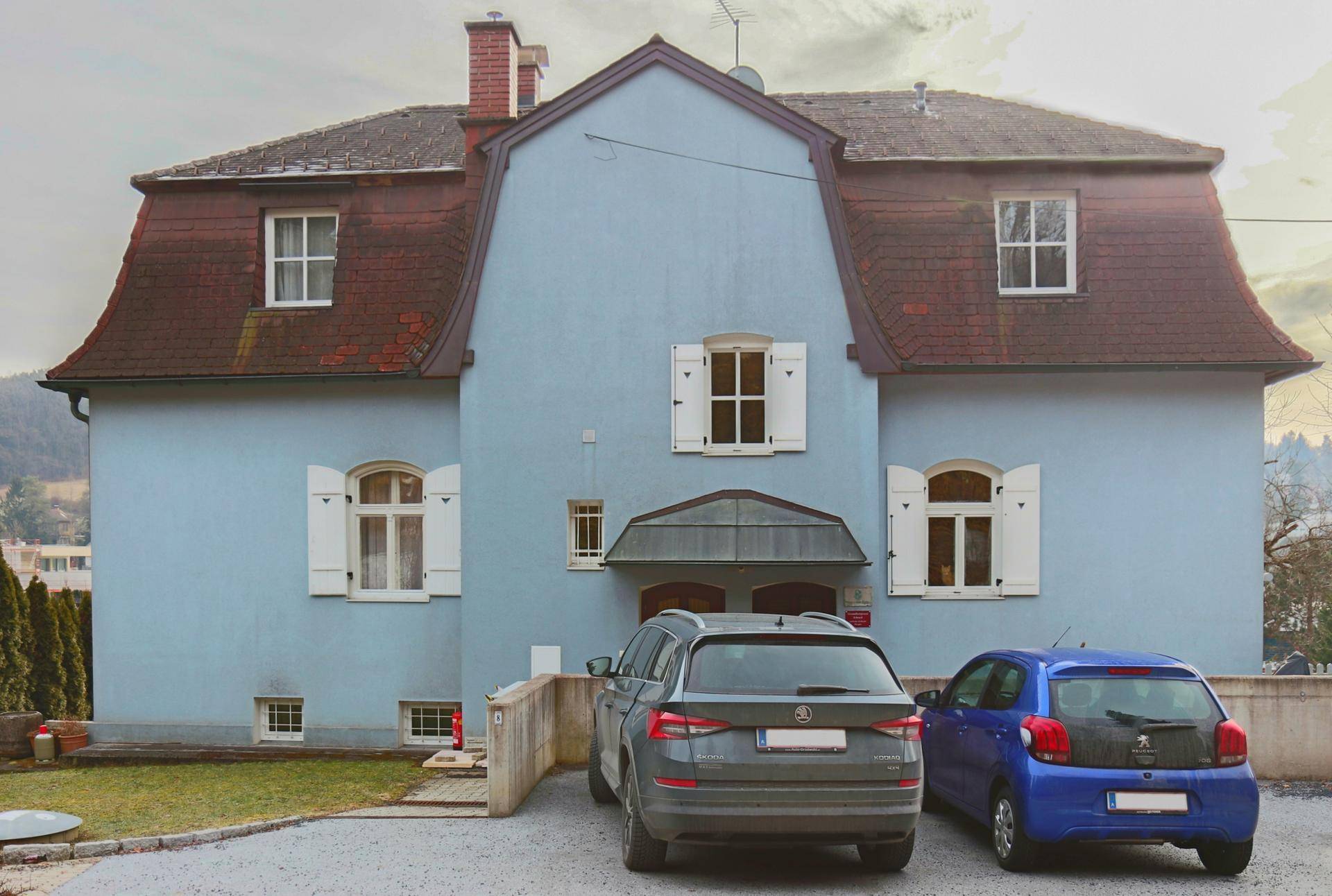 Historisches Gebäude mit blauer Fassade, Mansarddach und geparkten Autos vor dem Eingangsbereich.