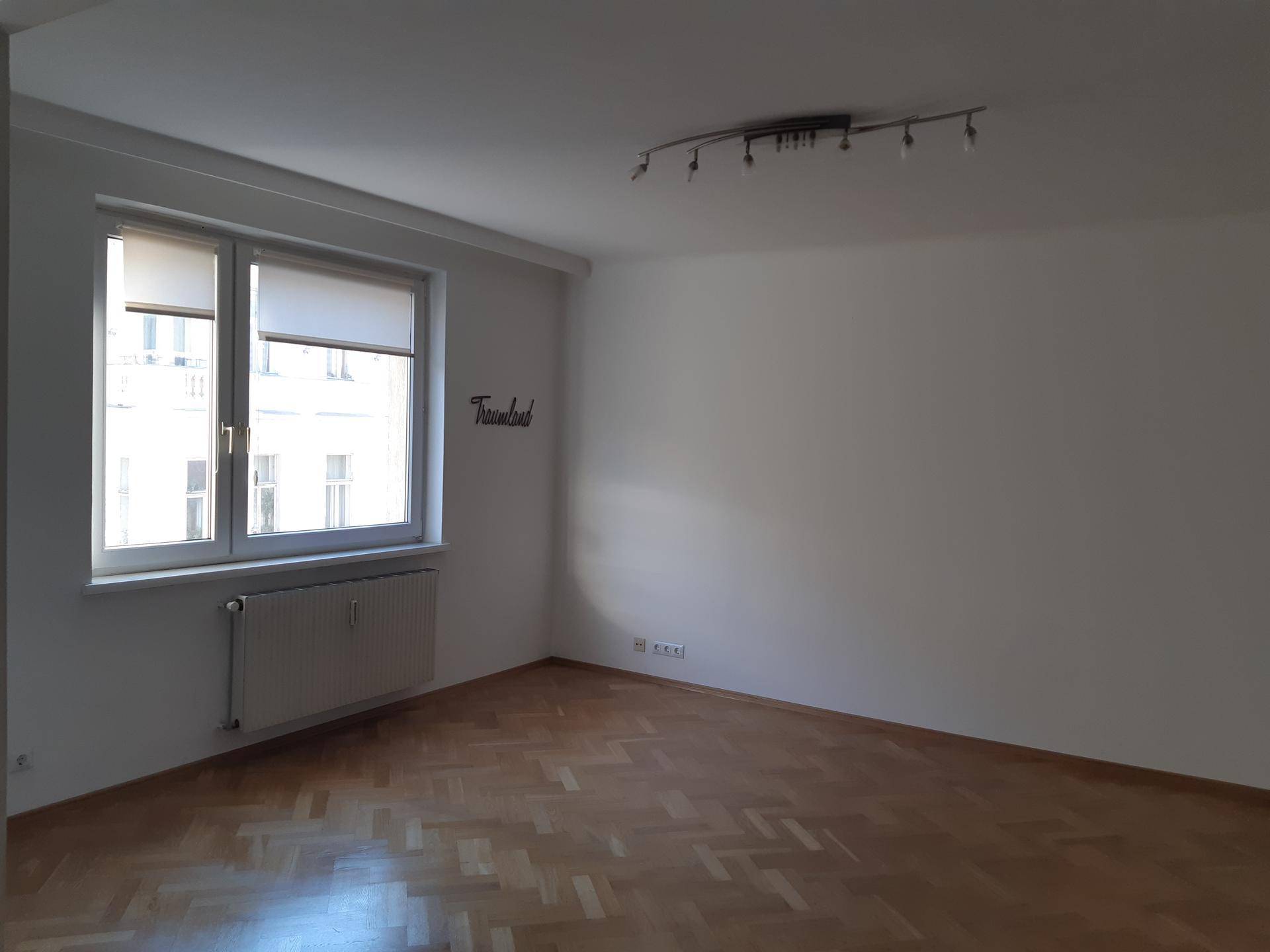 Leeres Wohnzimmer mit Fischgrätparkett, großem Fenster und Heizkörper.