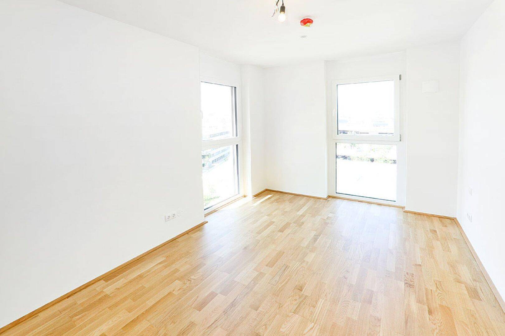 Helles, leeres Zimmer mit Parkettboden und zwei großen Fenstern.