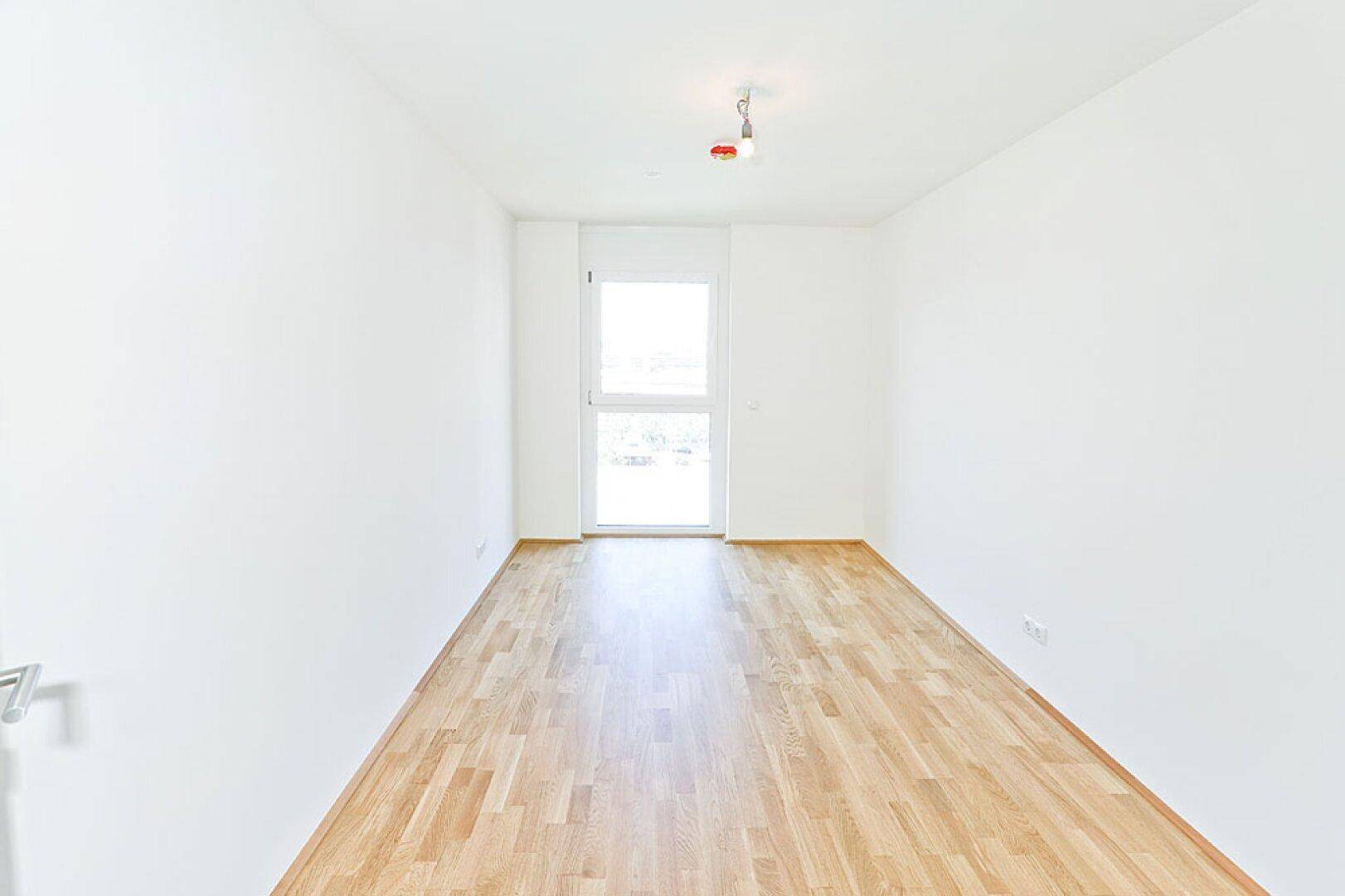 Leeres Zimmer mit Parkettboden und einem großen Fenster, das viel Licht hereinlässt.