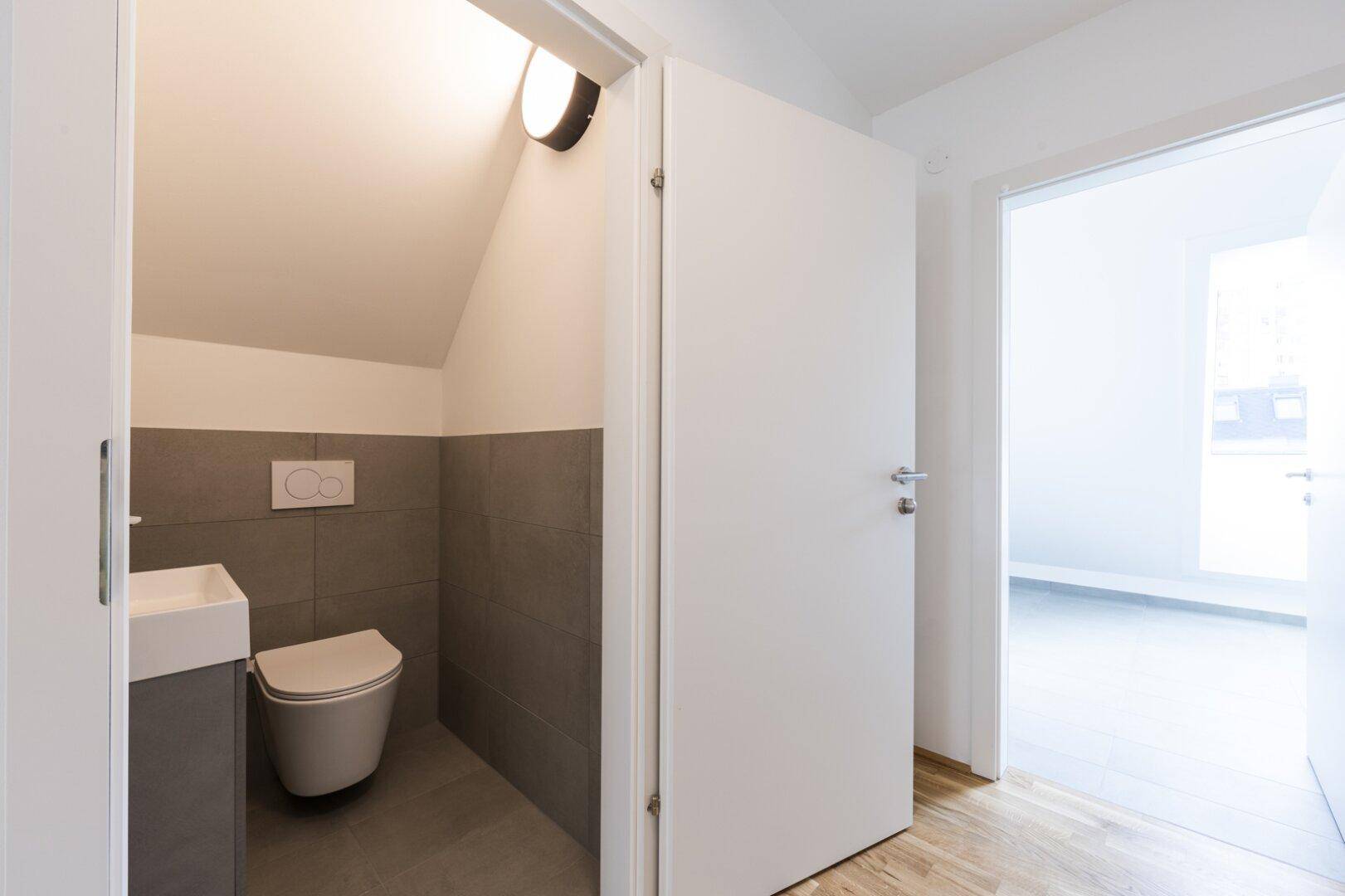 Modernes Gäste-WC mit grauen Fliesen, wandhängendem WC und kleinem Waschbecken.