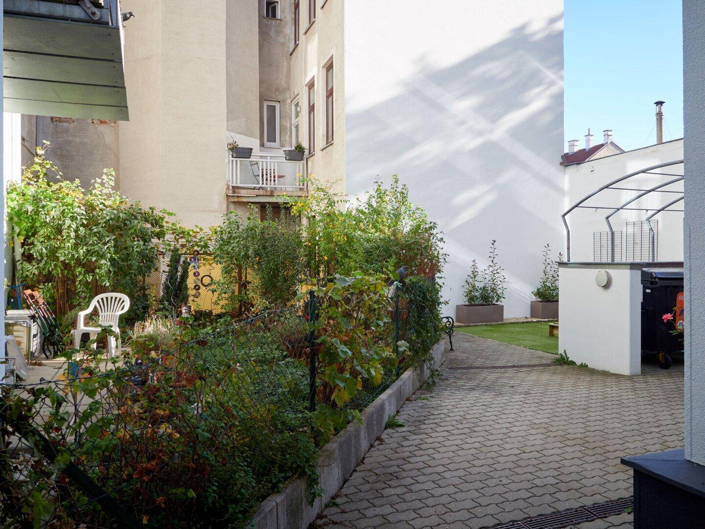 Grüner Innenhof mit gepflastertem Weg, üppiger Bepflanzung und einem kleinen Balkon im Hintergrund.