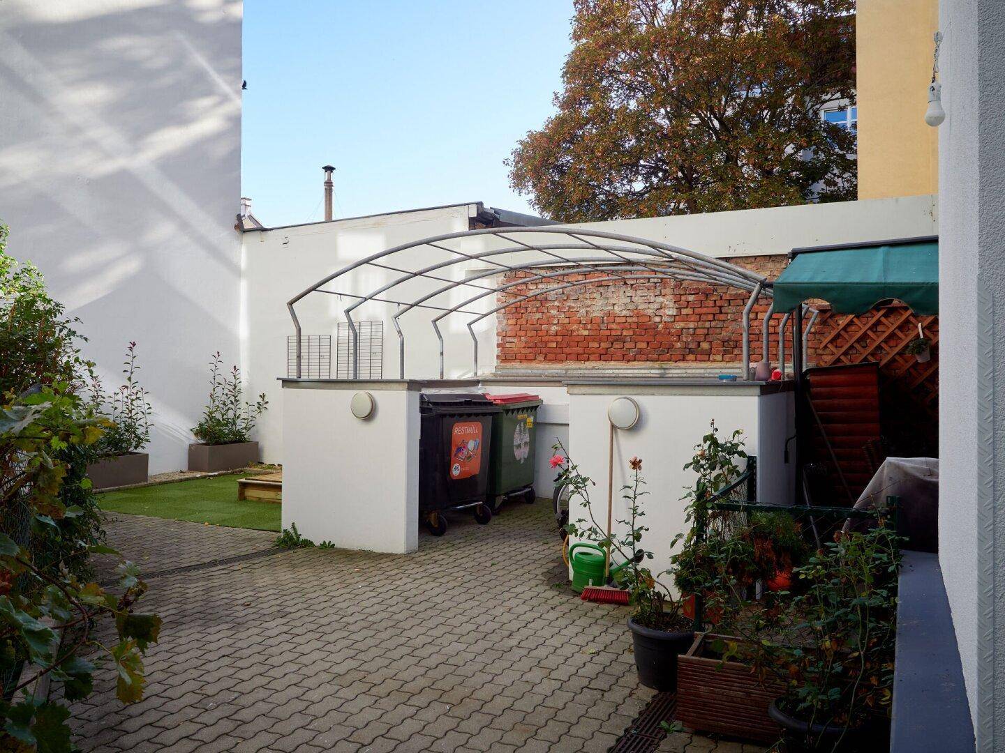 Innenhof mit gepflastertem Bereich, Mülltonnen und einer Metallkonstruktion im Hintergrund.