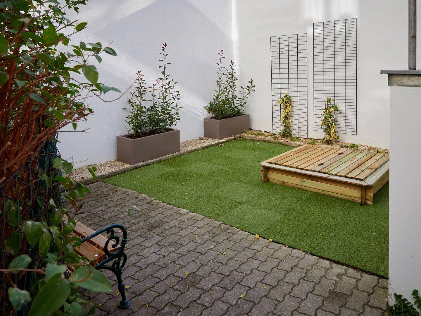 Kleiner Spielbereich im Innenhof mit Sandkasten, Kunstrasen und Pflanzkübeln.