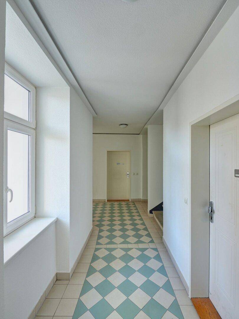 Heller Flur mit Fenster, kariertem Fliesenboden und weißen Wänden, der zu weiteren Räumen führt.