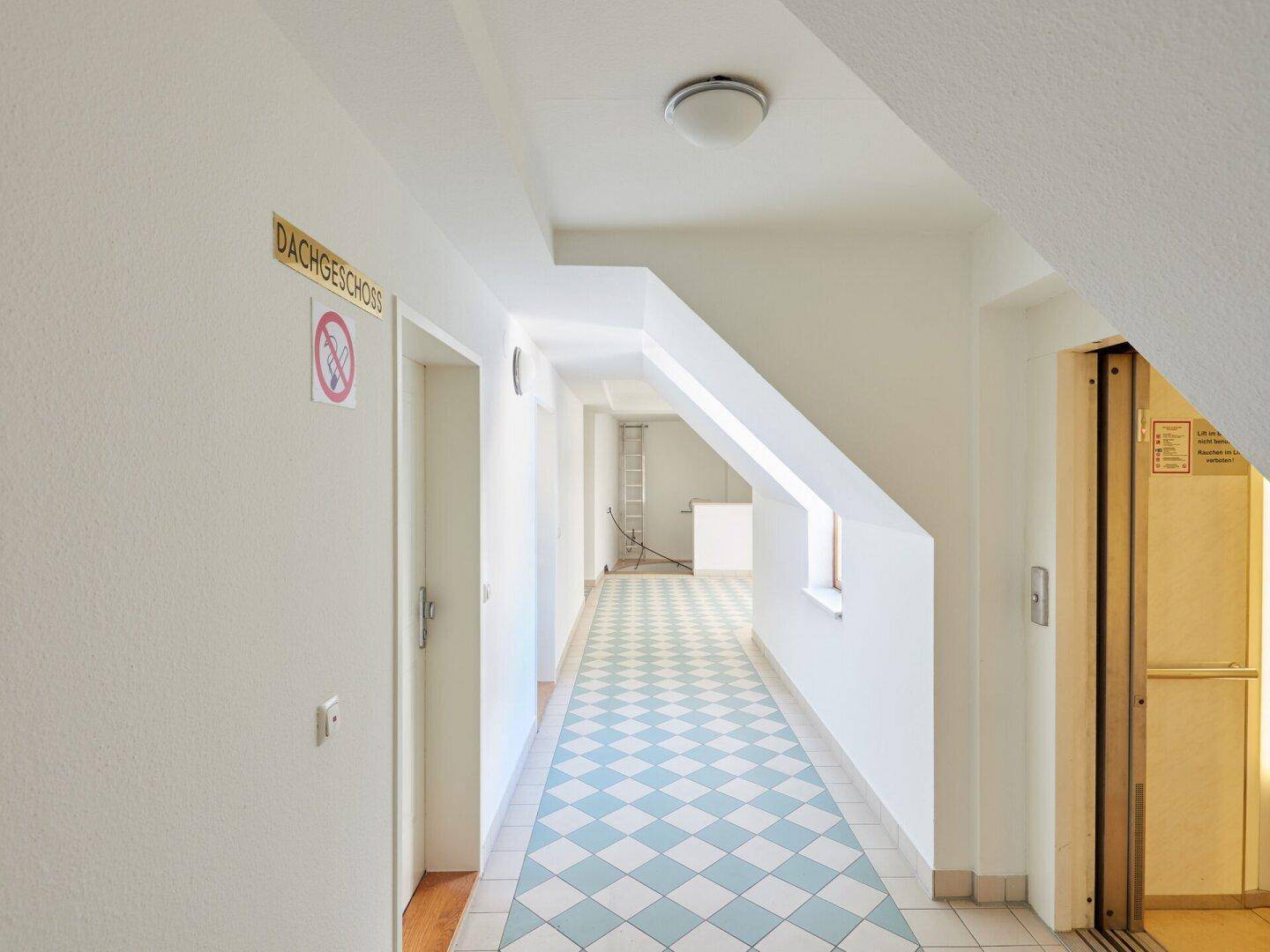 Heller Flur im Dachgeschoss mit schrägen Wänden, kariertem Boden und Aufzugzugang.
