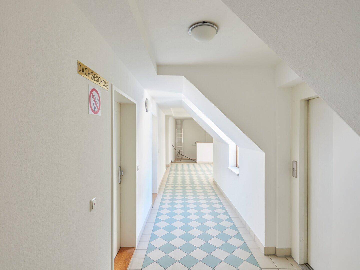 Heller Flur im Dachgeschoss mit schrägen Wänden, kariertem Boden und Aufzugzugang.