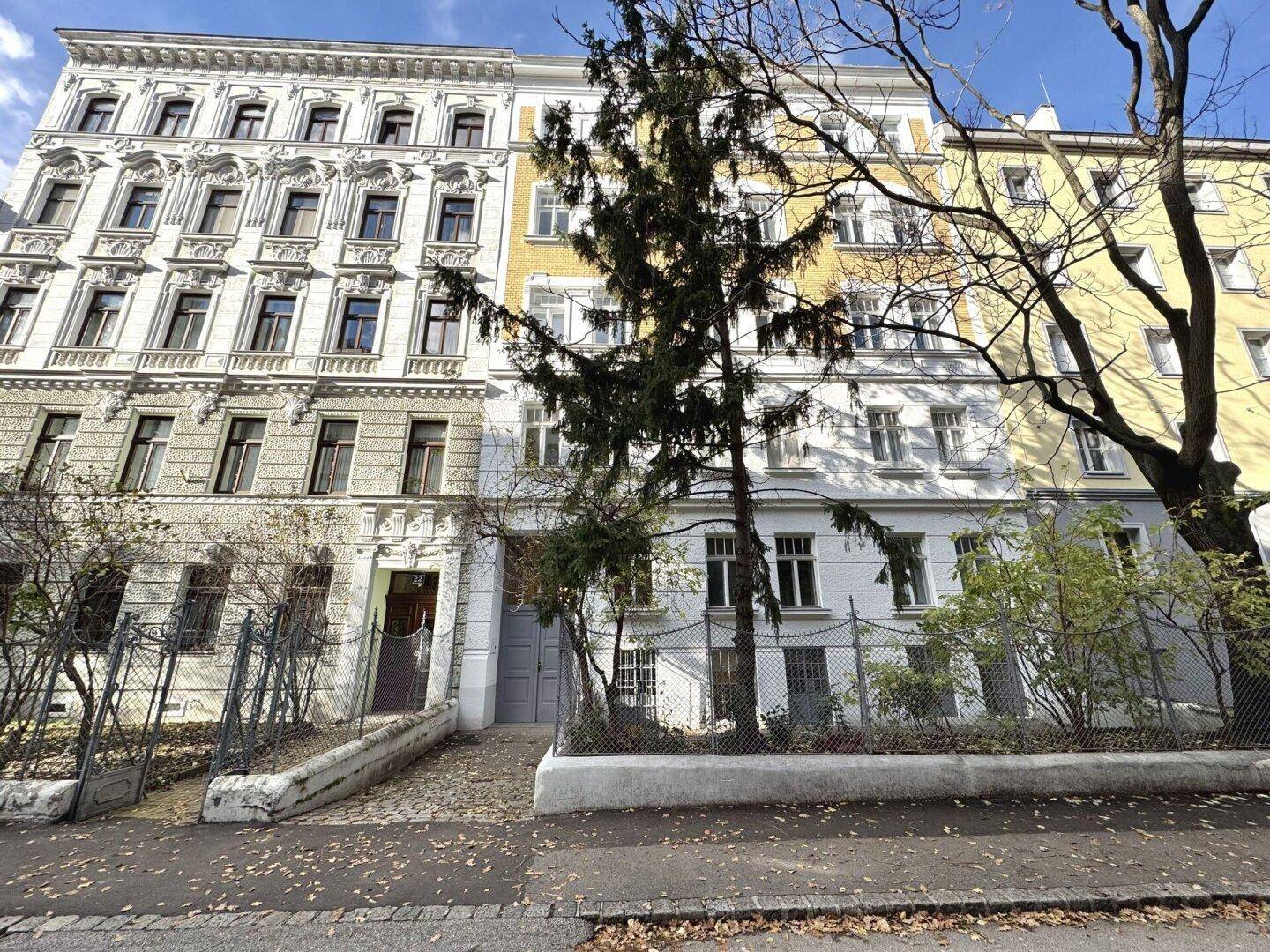 Gesamtansicht eines Altbaus mit gelber Fassade und umzäuntem Bereich davor.