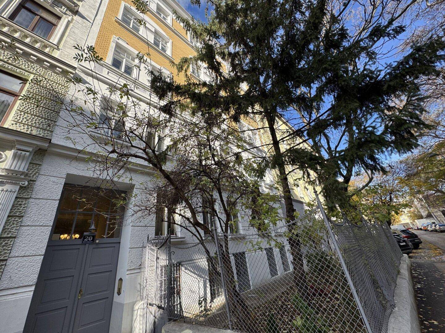 Seitenansicht eines Altbaus mit weißer Fassade und Bäumen im Vordergrund, an einer Straße gelegen.