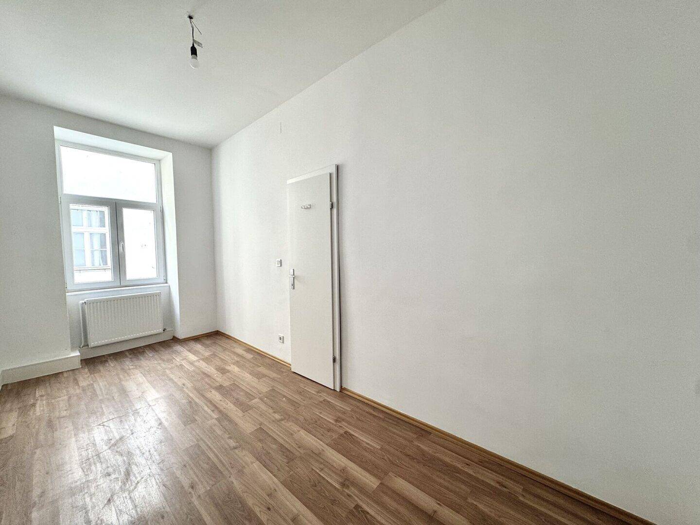 Helles Zimmer mit Holzboden und einem Fenster, bereit zur individuellen Einrichtung.