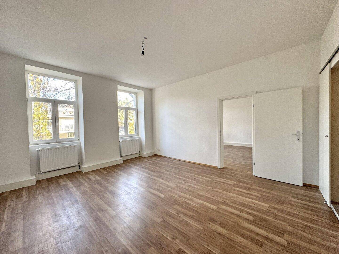 Großes Zimmer mit Holzboden und zwei Fenstern, mit Zugang zu einem weiteren Raum.