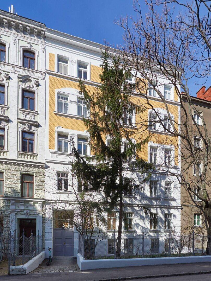 Historisches Mehrfamilienhaus mit gelber Fassade und grünen Bäumen davor.