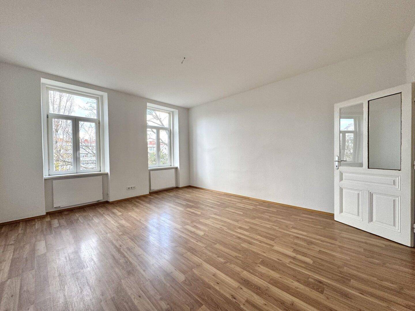 Helles, leeres Zimmer mit Laminatboden und zwei großen Fenstern, die viel Licht hereinlassen.