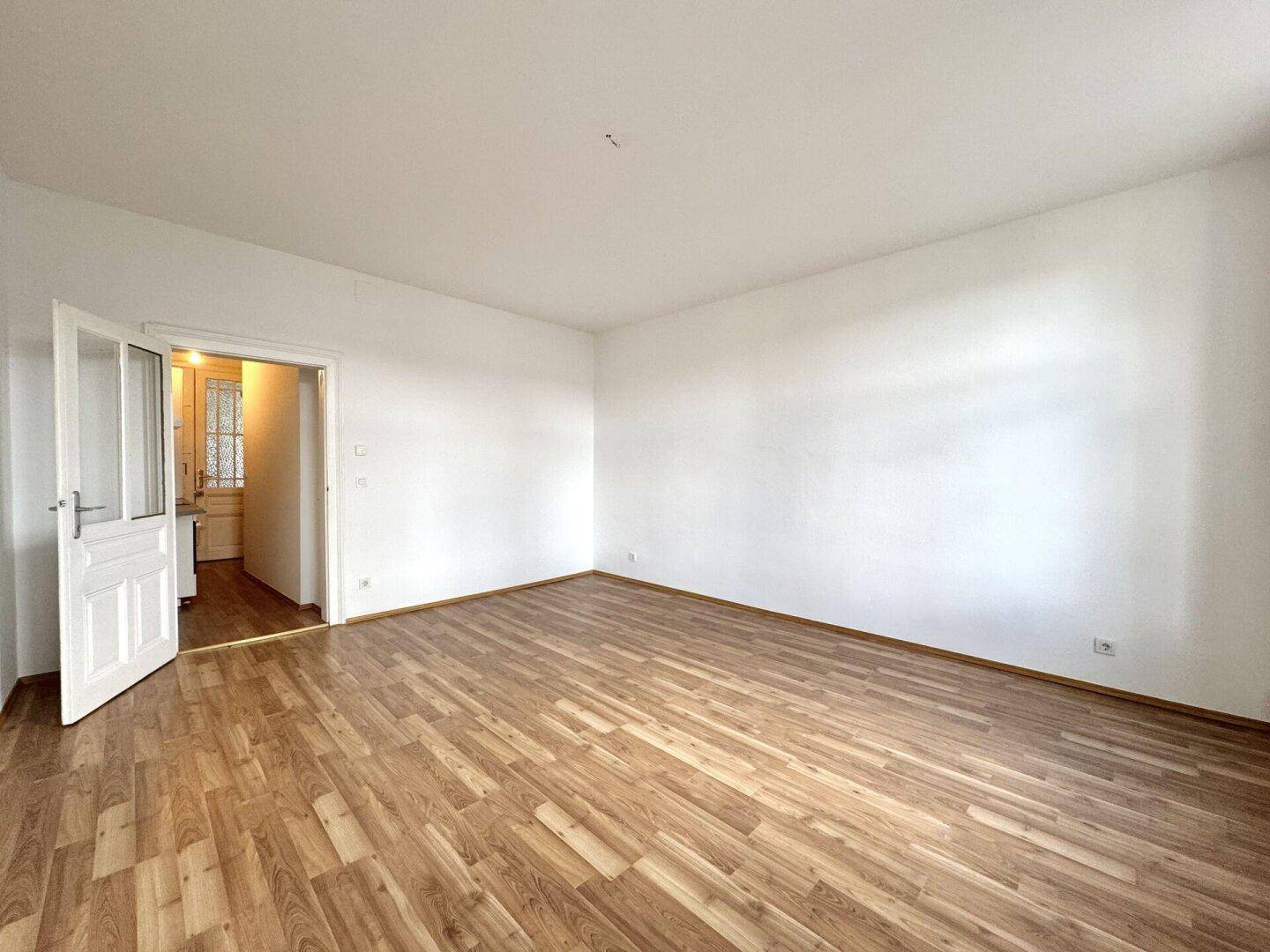Geräumiges, leeres Zimmer mit Laminatboden und weiß gestrichenen Wänden, mit Zugang zur Küche.
