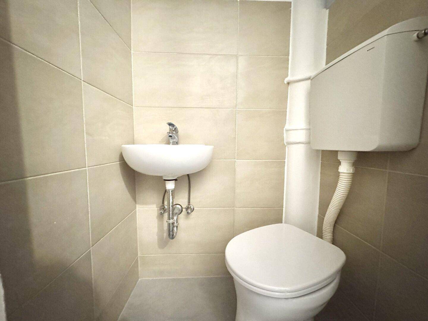Kleines WC mit beigefarbenen Fliesen, wandmontierter Toilette und kompaktem Waschbecken.