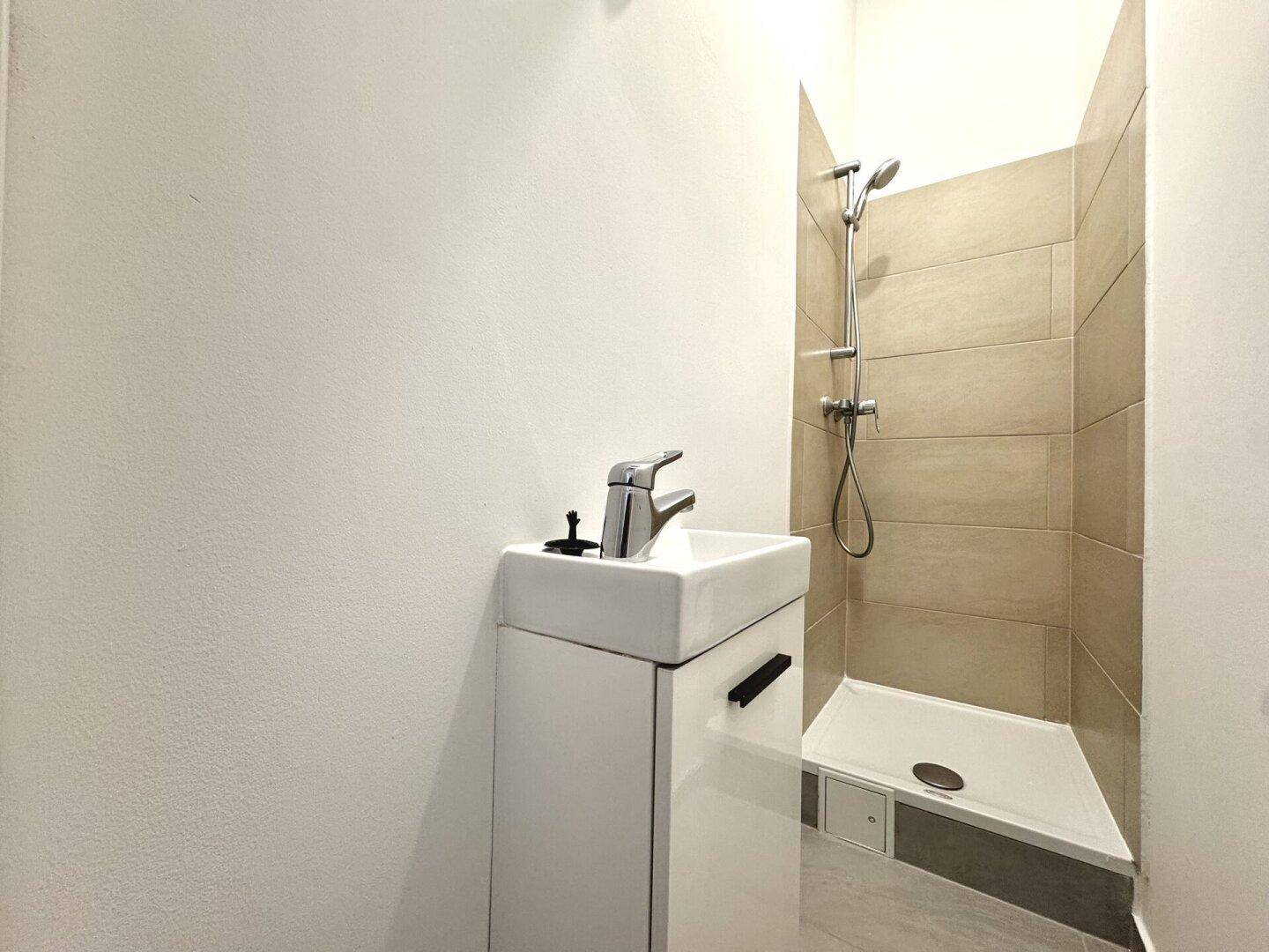 Modernes Badezimmer mit begehbarer Dusche, beigefarbenen Fliesen und kleinem Waschtisch.