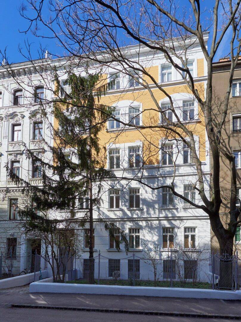 Historisches Gebäude mit weißer und gelber Fassade, umgeben von Bäumen unter blauem Himmel.