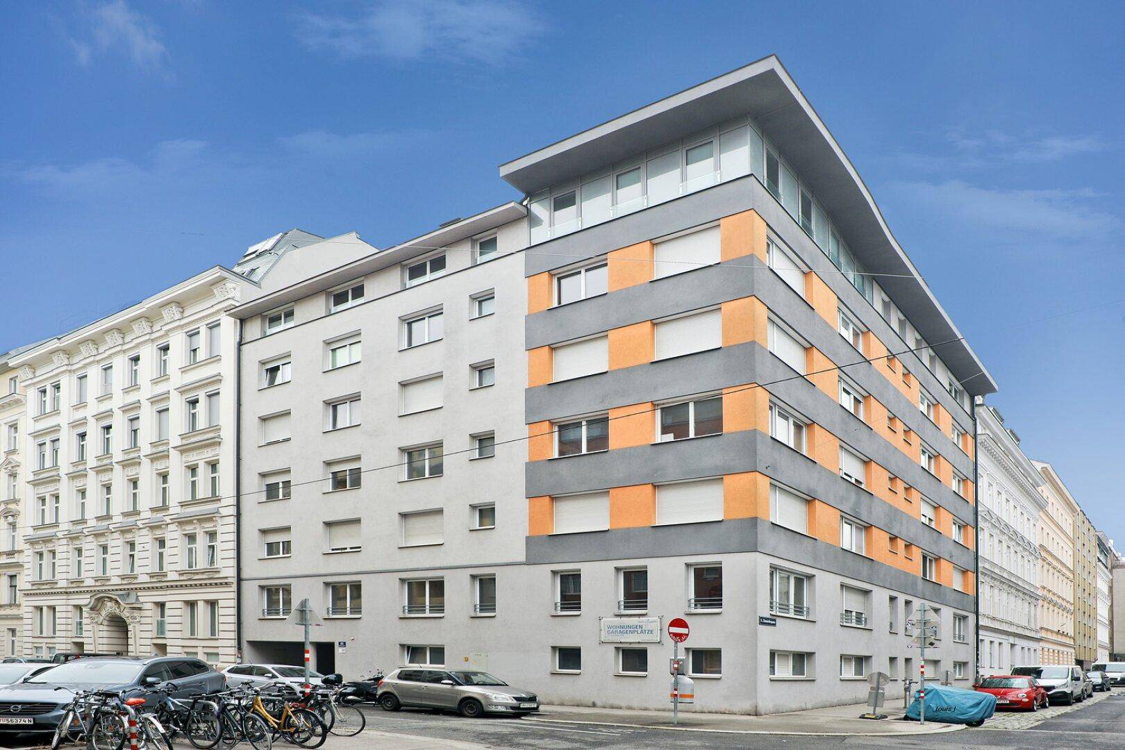 Modernes Mehrfamilienhaus mit grauer und orangefarbener Fassade an einer städtischen Straße gelegen.
