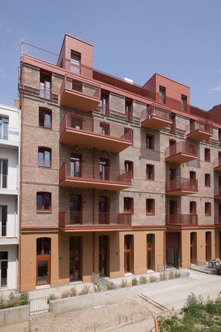 Backsteingebäude mit zahlreichen Balkonen und roten Fensterrahmen unter blauem Himmel.