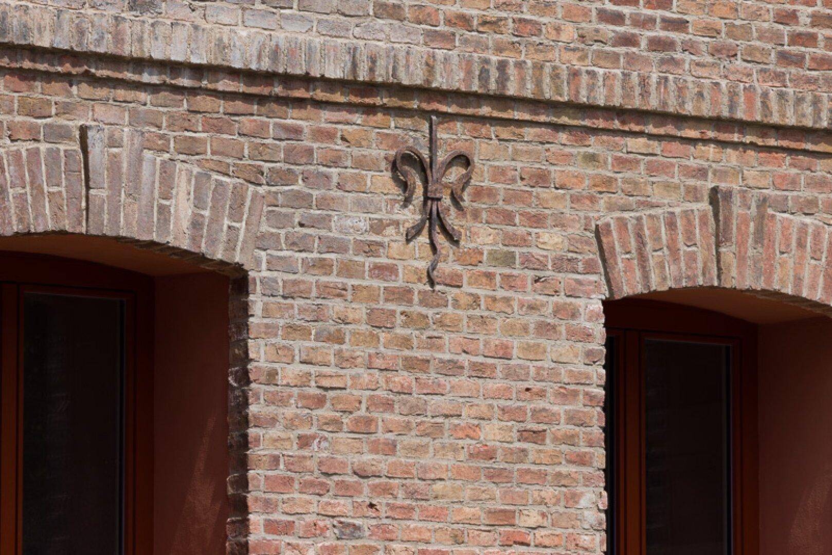 Detailansicht einer Backsteinwand mit verzierten Ziegelbögen und einem schmiedeeisernen Ornament.