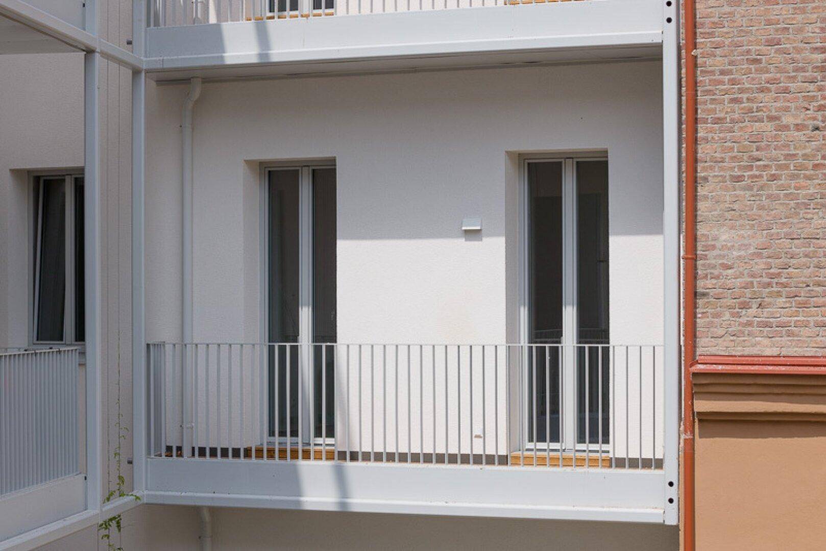 Weißer Balkon an einer modernen Fassade mit großen Fenstertüren und Metallgeländer.