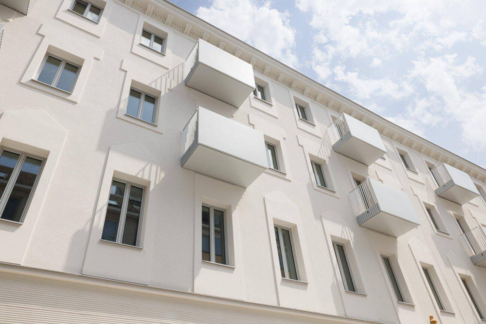 Moderne weiße Fassade mit mehreren Balkonen und großen Fenstern, unter blauem Himmel.