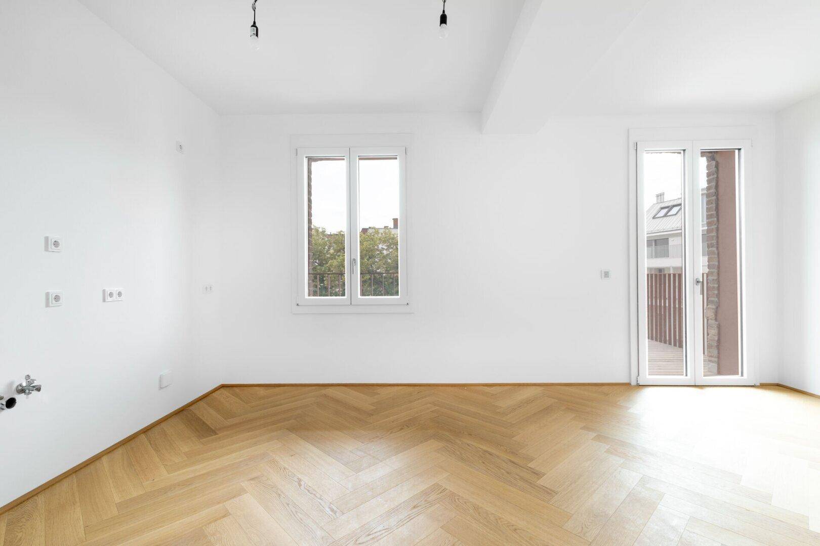 Geräumiger, heller Raum mit Fischgrätparkettboden, weißen Wänden und Zugang zum Balkon.