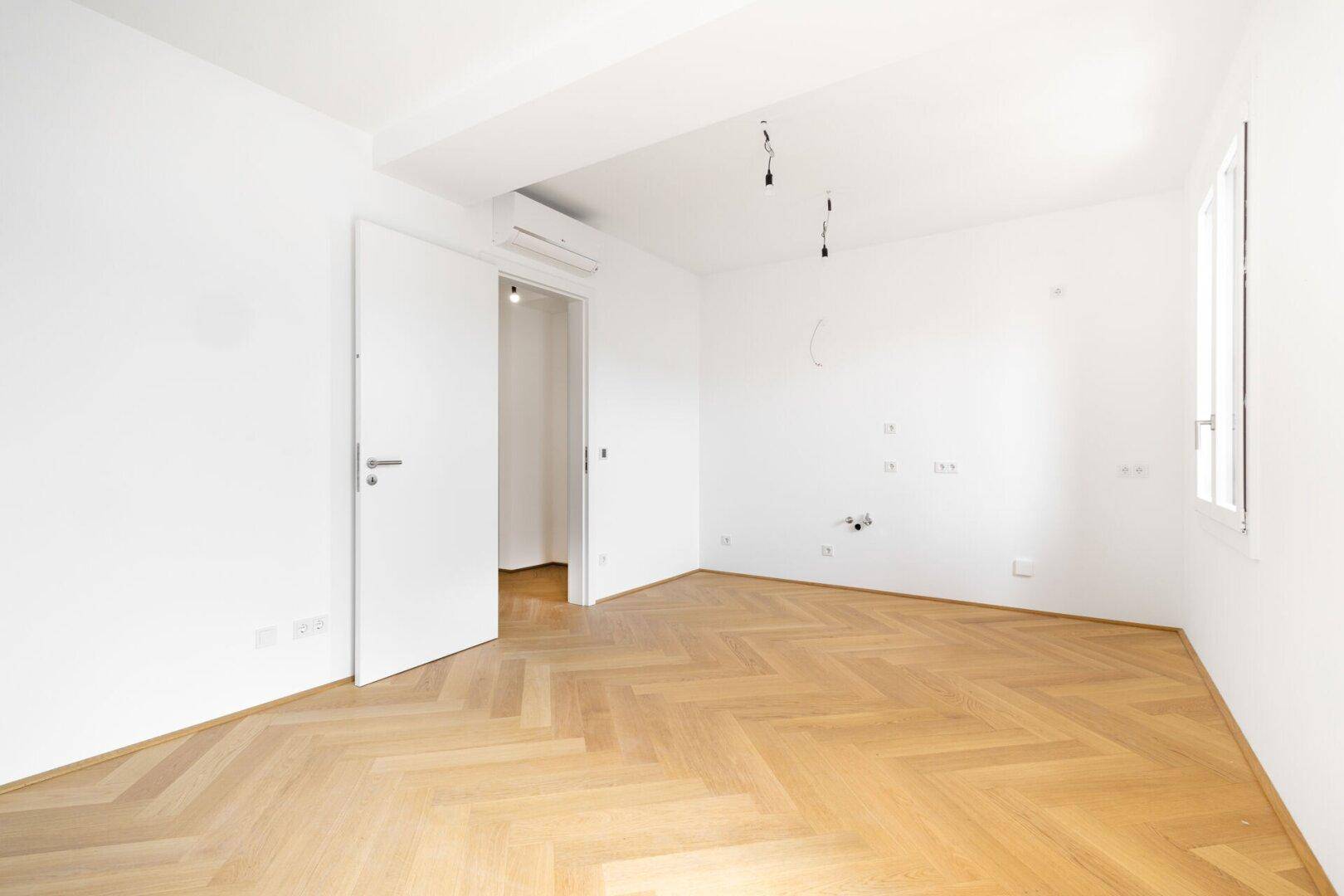 Leerer, heller Raum mit Fischgrätparkettboden, weißen Wänden und einem großen Fenster.