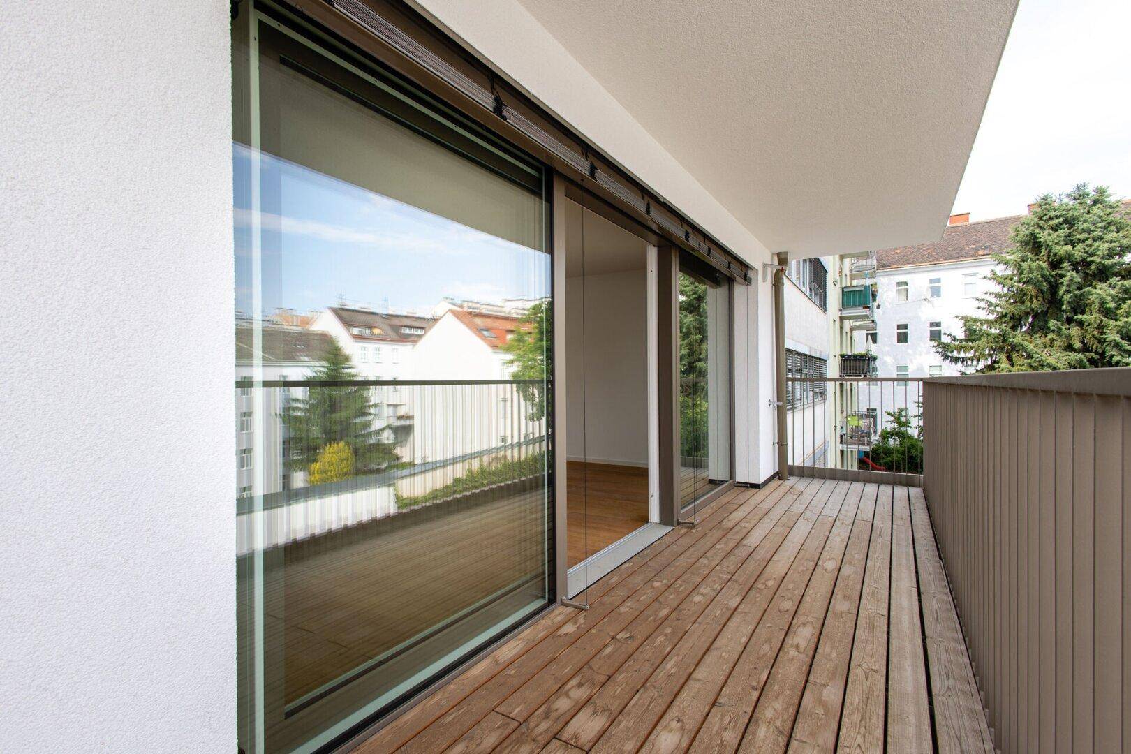 Geräumiger Balkon mit Holzboden und Blick auf die umliegenden Gebäude und Bäume.