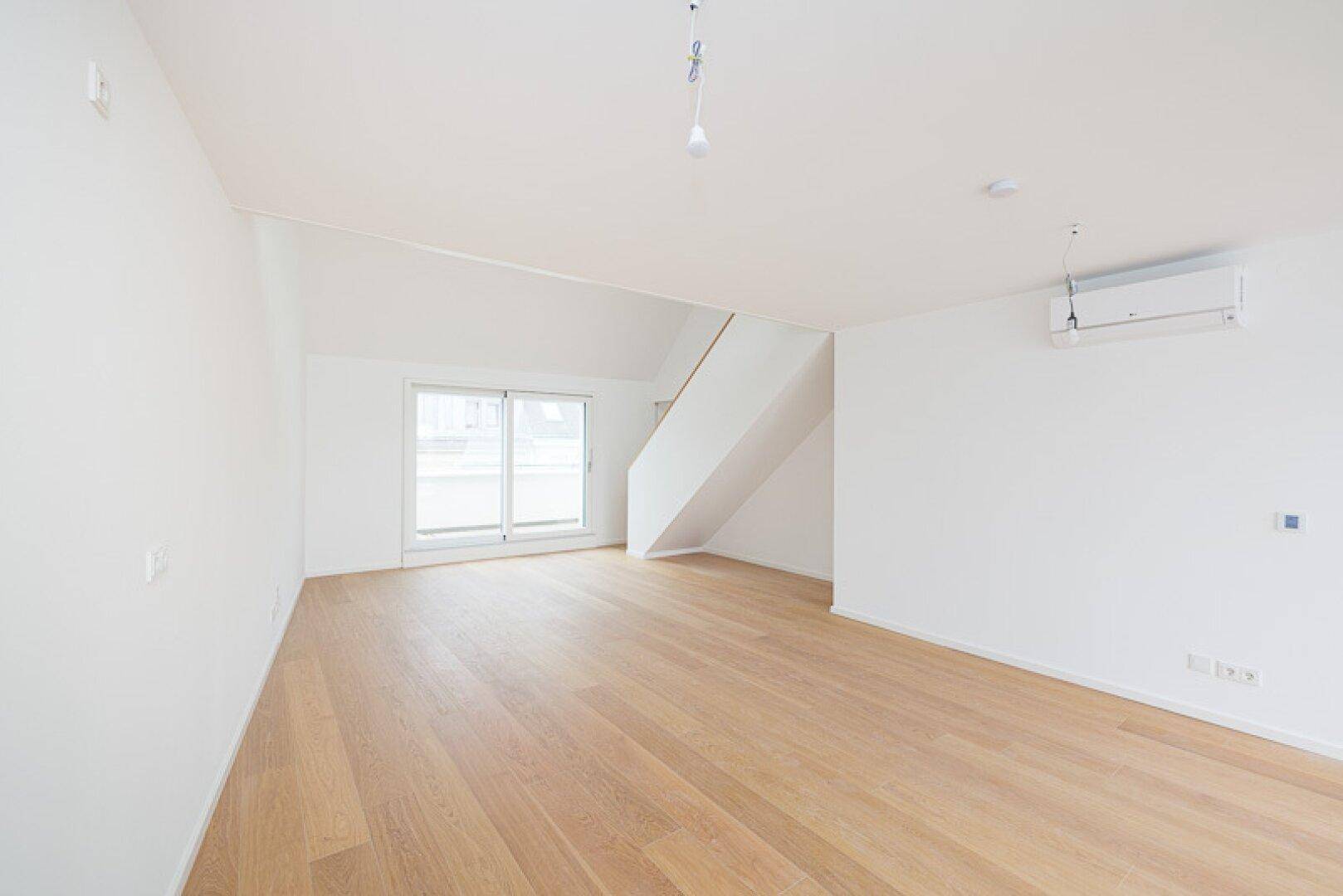 Geräumiger Wohnbereich mit Holzparkett, weiß gestrichenen Wänden und Zugang zu einem Balkon.