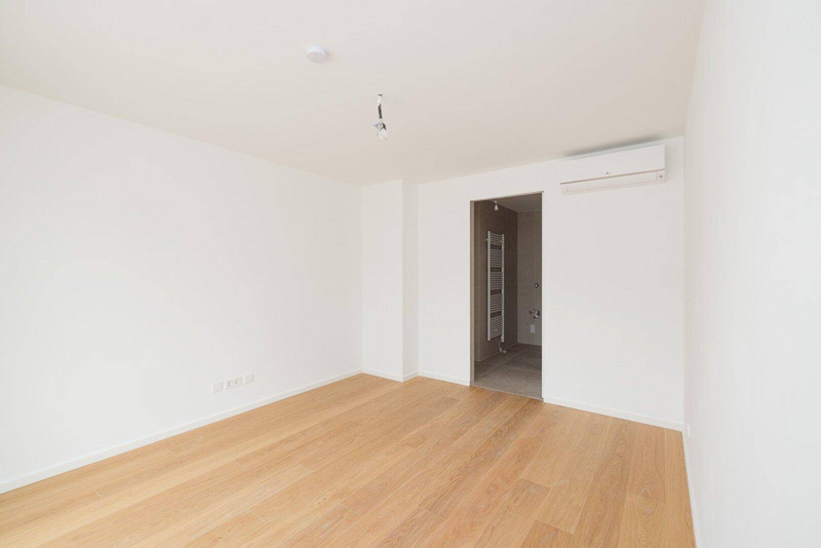 Helles Schlafzimmer mit Holzparkett, Klimaanlage und direktem Zugang zu einem Badezimmer.