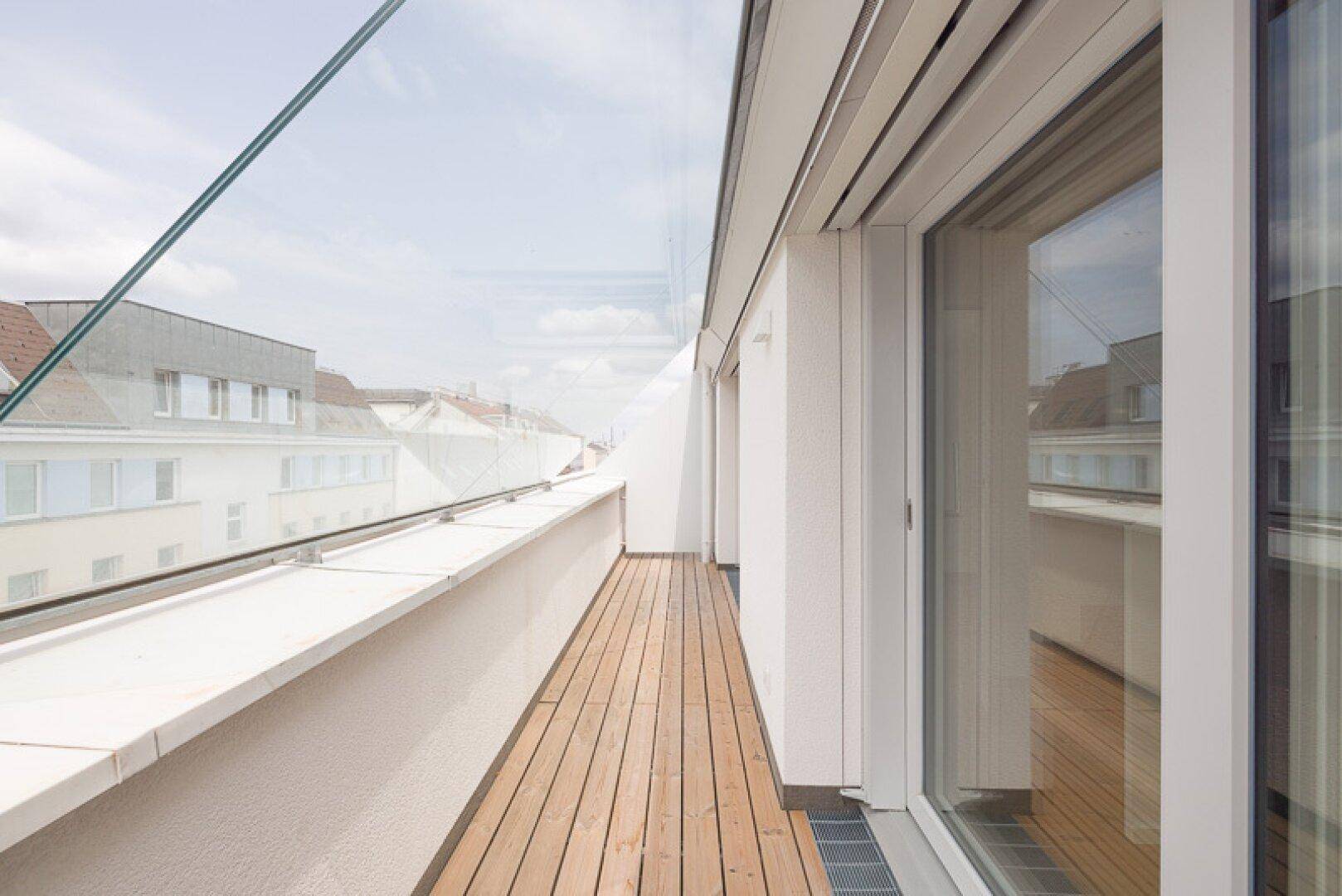 Langer, schmaler Balkon mit Holzboden und Glasgeländer, der einen seitlichen Blick bietet.