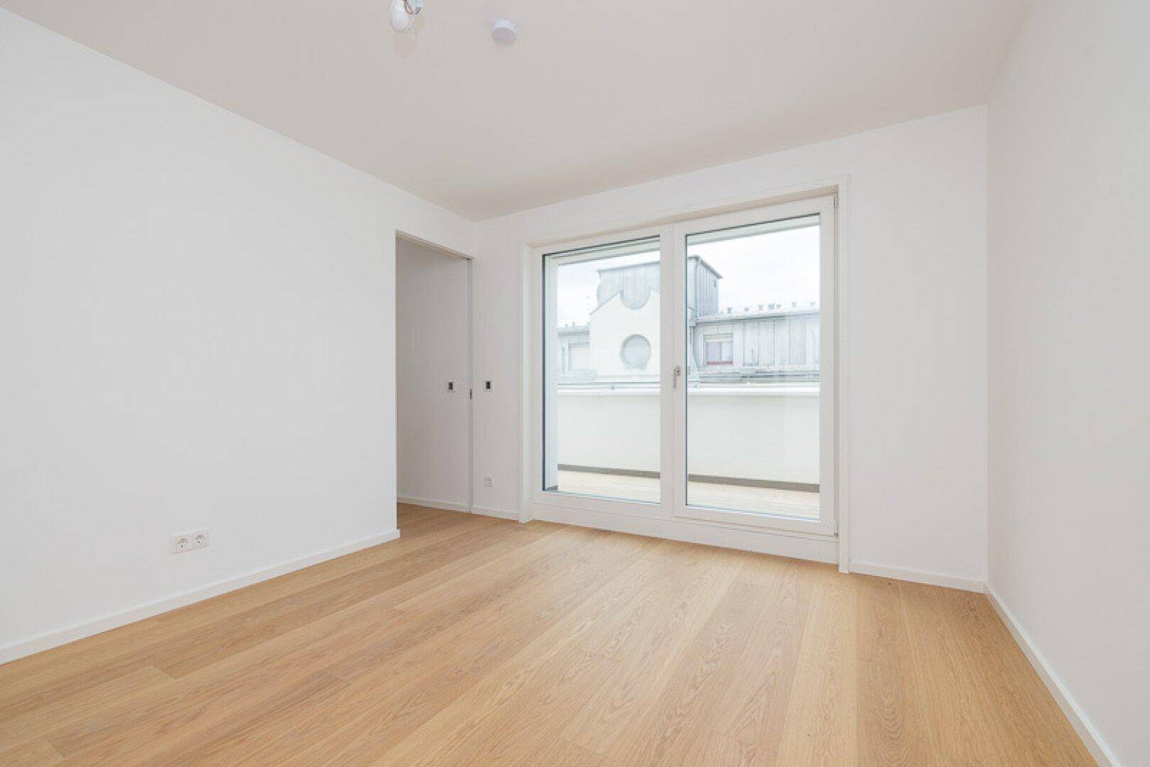 Geräumiges Schlafzimmer mit Holzparkett und großen Fenstern, die Zugang zu einem Balkon bieten.