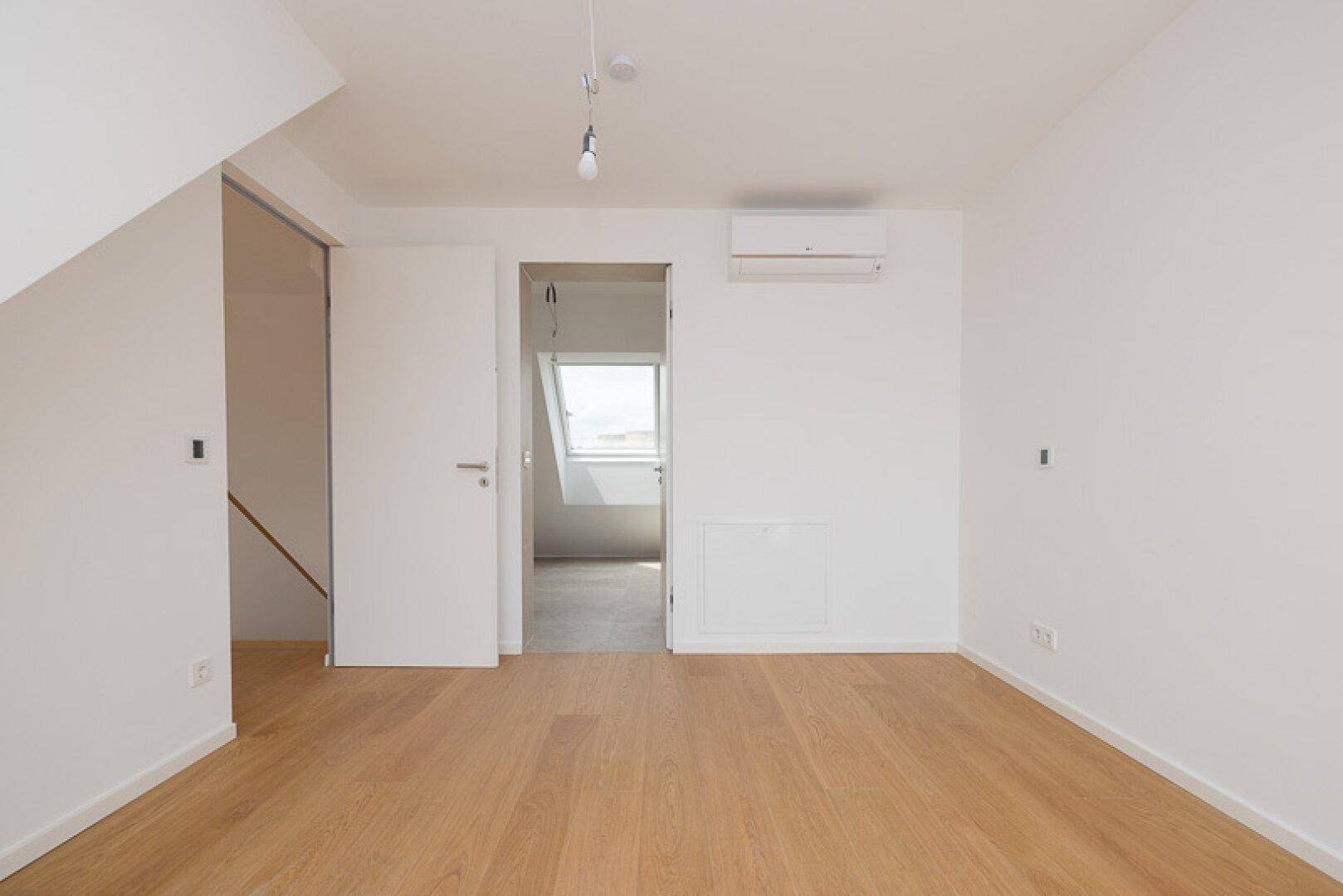 Geräumiges Schlafzimmer mit Holzparkett, Klimaanlage und Zugang zu einem weiteren Raum.