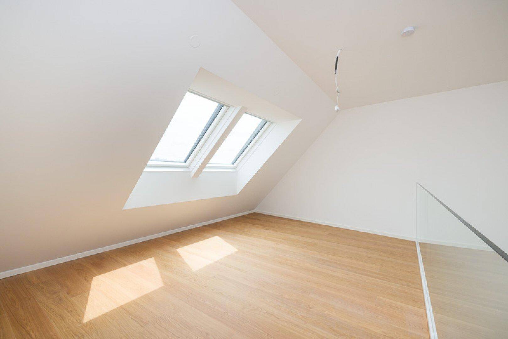 Helles Zimmer im Dachgeschoss mit Holzparkett, schrägen Wänden und zwei Dachfenstern.