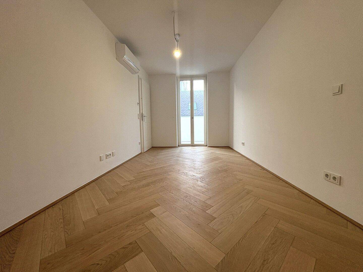 Helles Zimmer mit Fischgrätparkett und Zugang zu einem Balkon oder einer Terrasse.