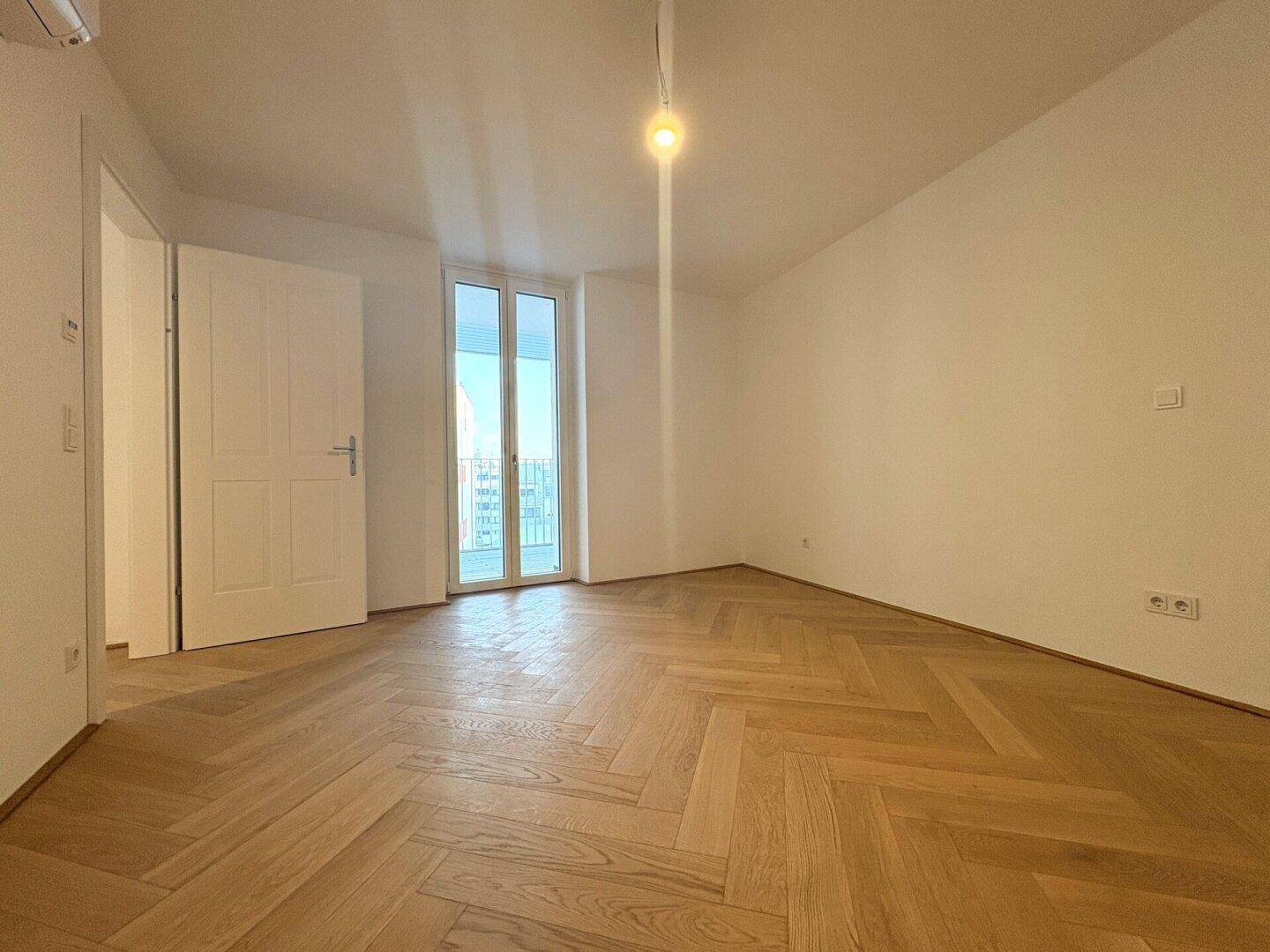 Helles Zimmer mit Fischgrätparkett und Zugang zu einem Balkon oder einer Terrasse.