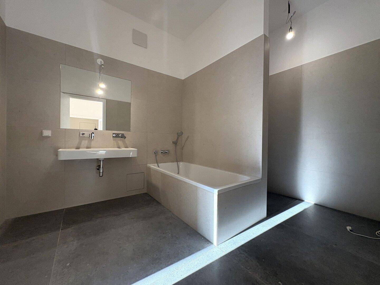 Modernes Badezimmer mit beigefarbenen Fliesen, Badewanne und dunklem Boden.