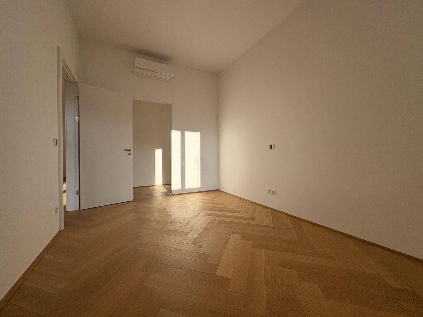 Geräumiges Schlafzimmer mit Fischgrätparkett, weißen Wänden und viel Tageslicht.