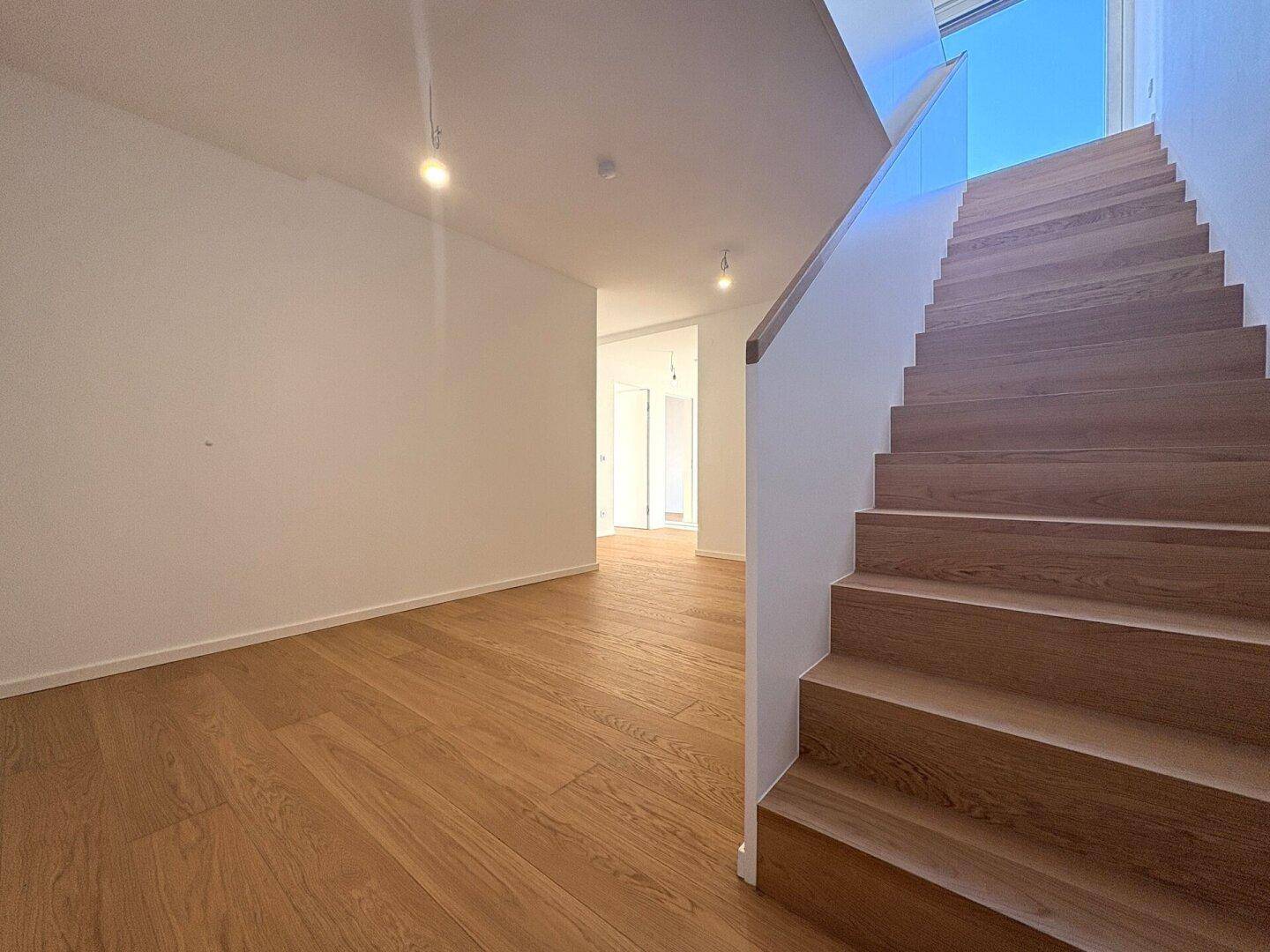 Heller Flur mit modernem Holzparkett und einer eleganten Treppe mit Glasgeländer.