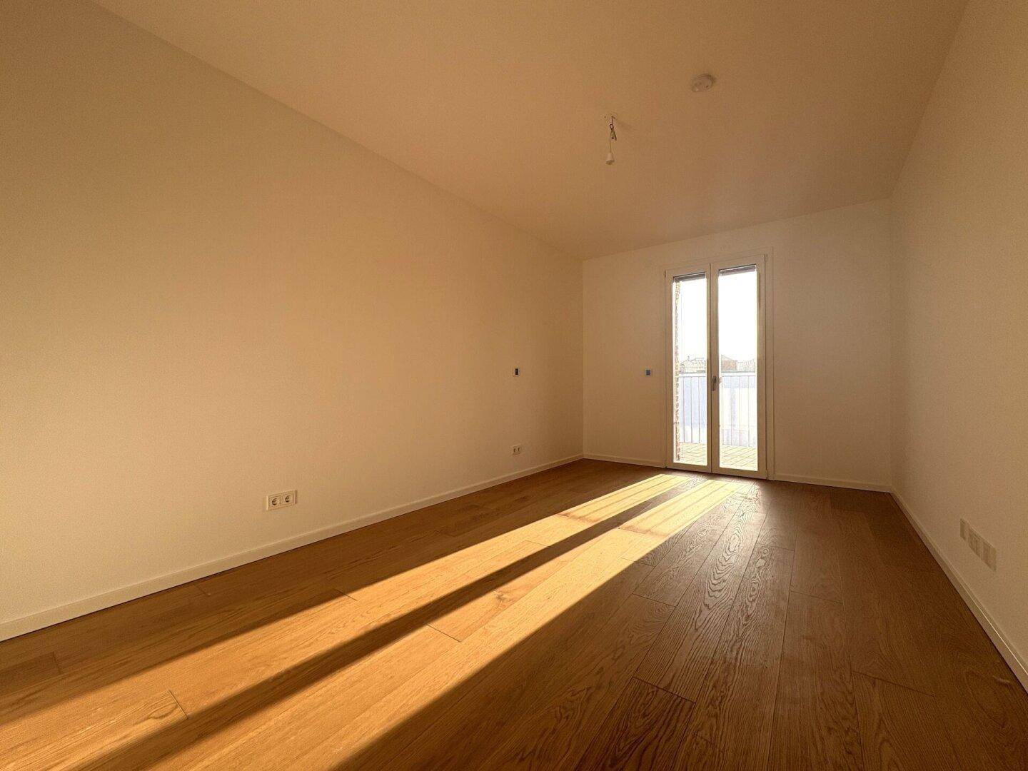 Helles Zimmer mit Holzparkett und Zugang zu einem Balkon oder einer Terrasse, durchflutet von Sonnenlicht.