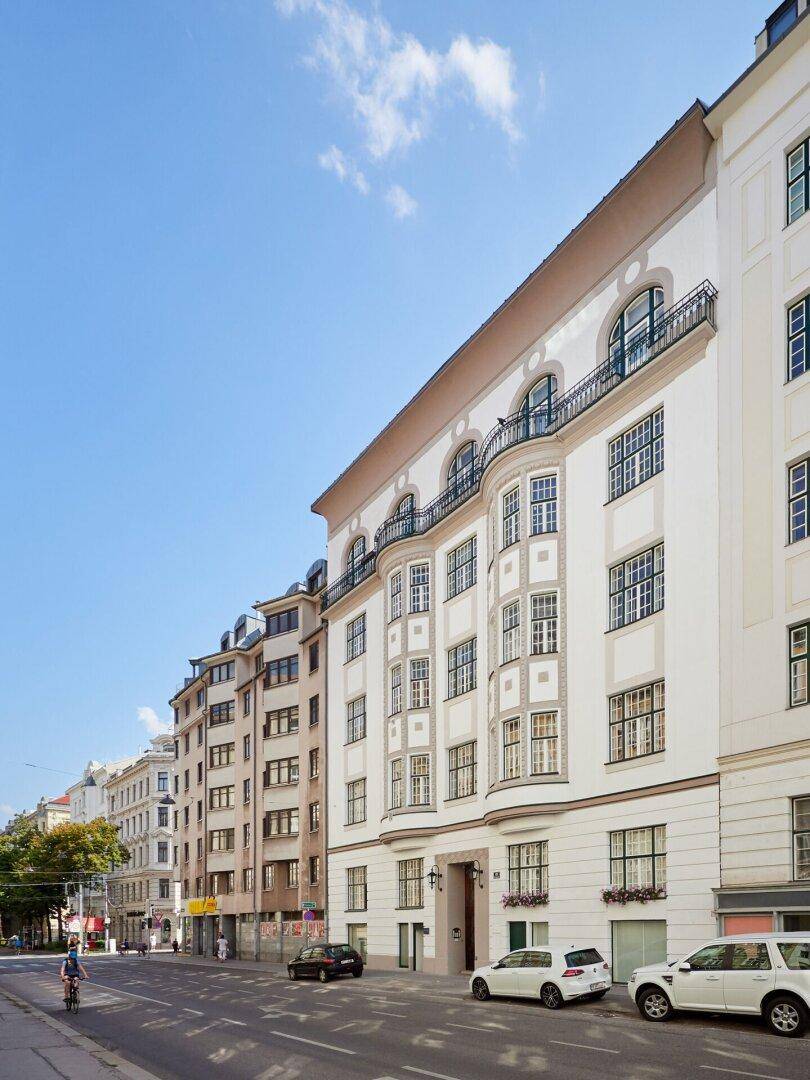 Historisches Wohngebäude an einer städtischen Straße mit geparkten Autos und blauem Himmel.