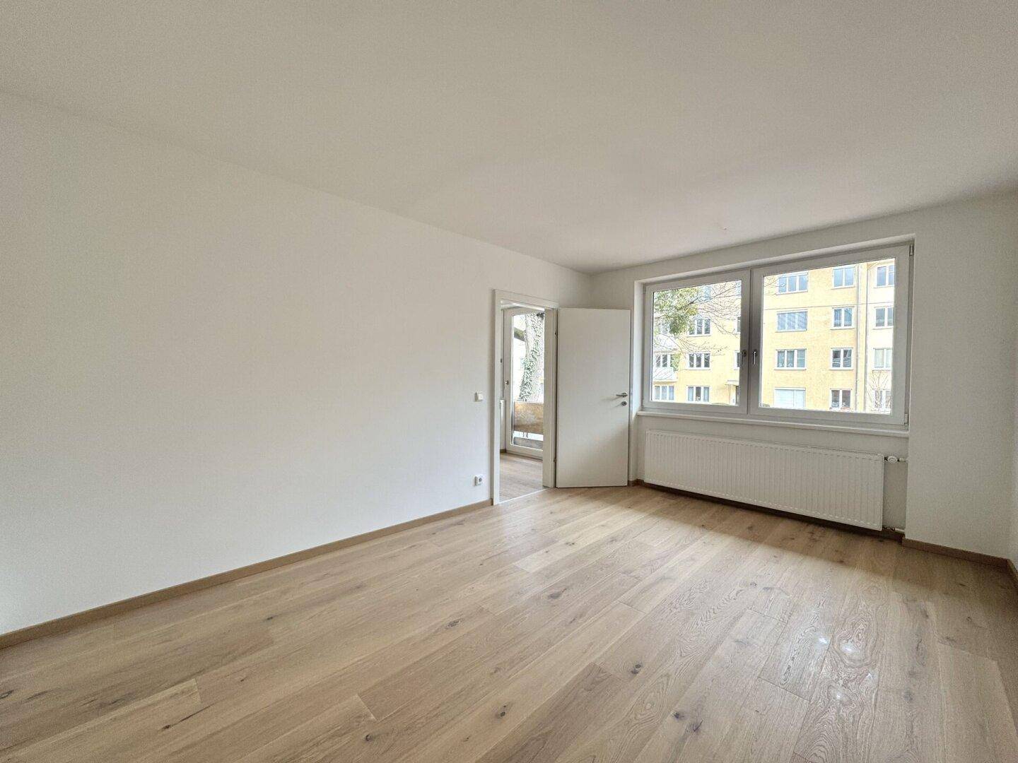 Geräumiges Zimmer mit Parkettboden, Fenster und Tür zu einem weiteren Raum.
