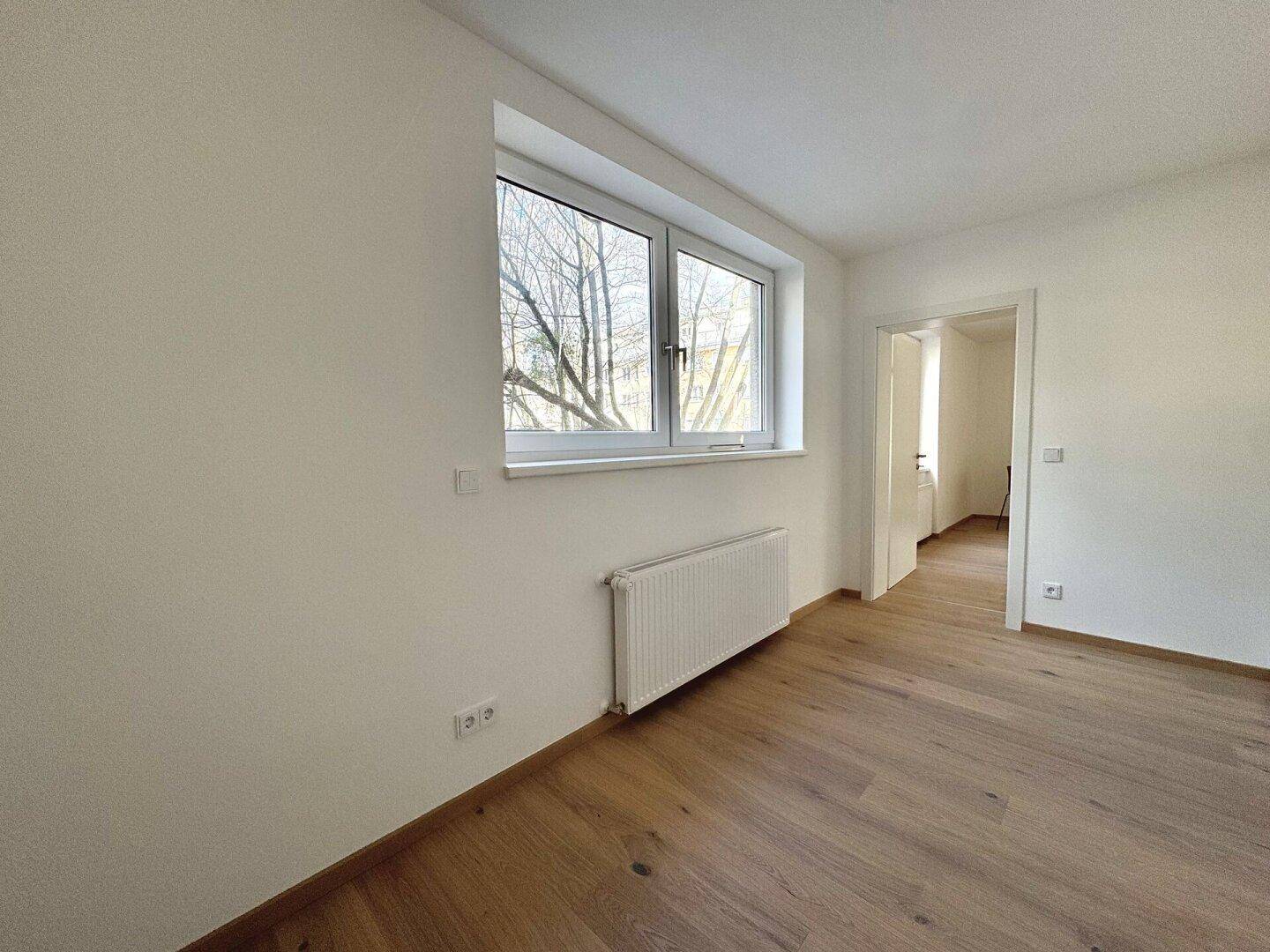 Geräumiges Zimmer mit Parkettboden, Fenster und Zugang zu einem weiteren Raum.
