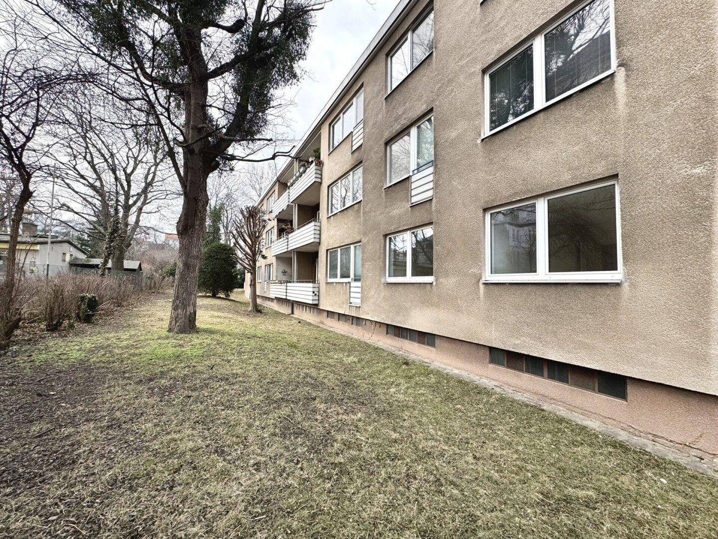 Seitenansicht eines Mehrfamilienhauses mit Balkonen und Bäumen im Vordergrund.