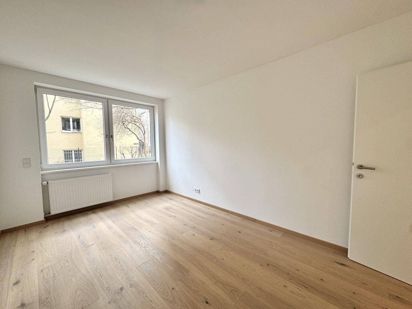 Geräumiges Zimmer mit Parkettboden, Fenster und Tür zu einem weiteren Raum.