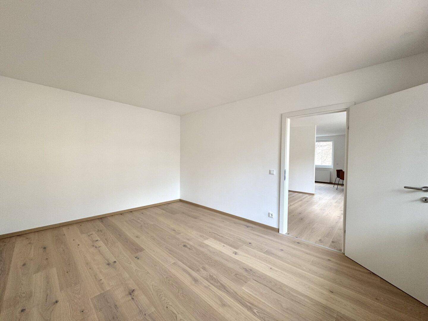 Leerer Raum mit Parkettboden und weißen Wänden, der durch eine Tür mit einem weiteren Zimmer verbunden ist.