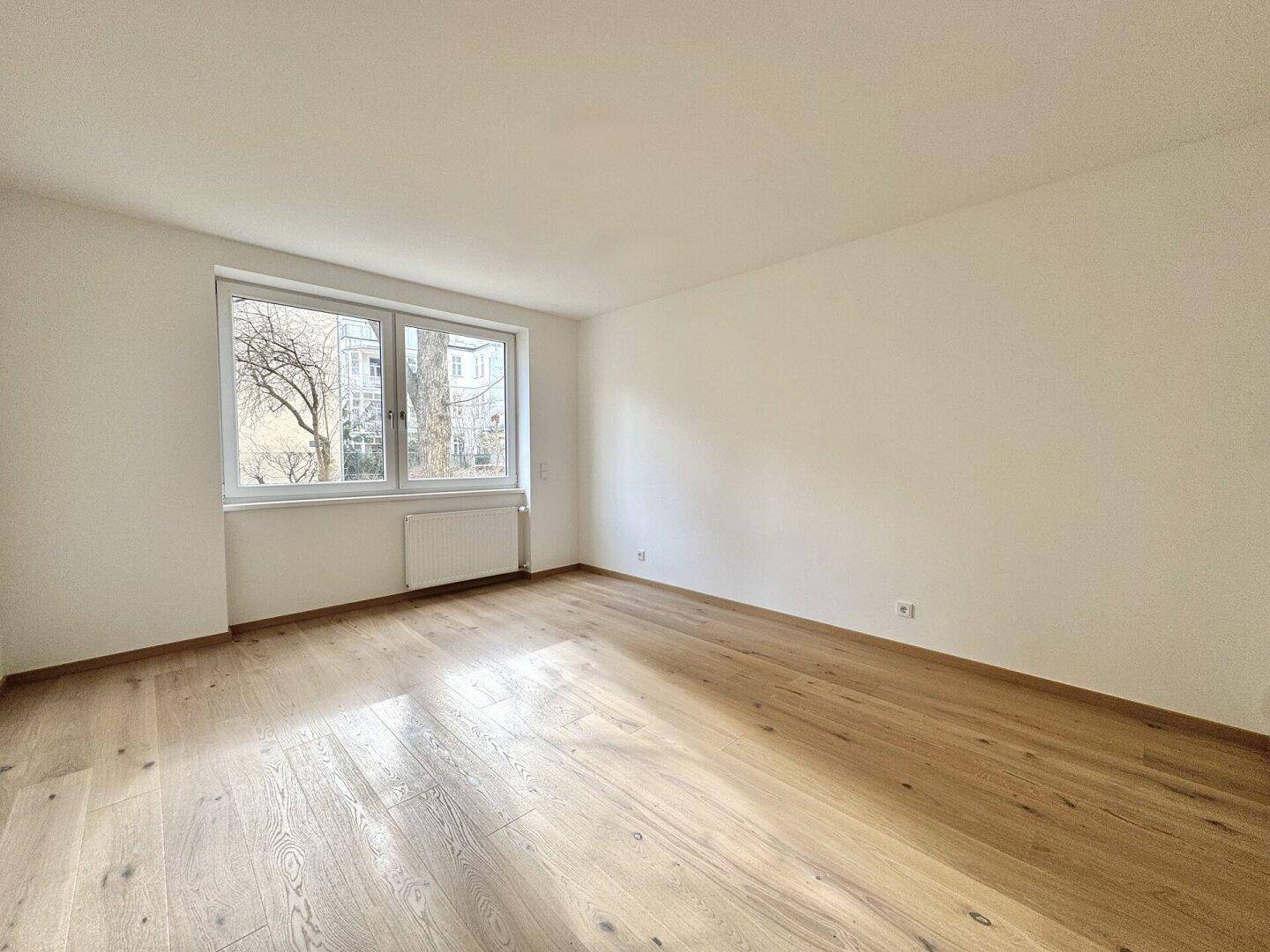 Geräumiger, heller Raum mit großem Fenster und Holzparkettboden, ideal für Wohn- oder Schlafbereich.