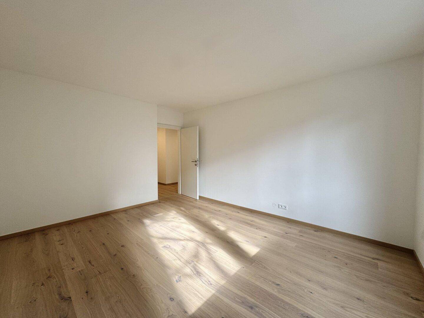 Heller Raum mit Holzparkettboden und offener Tür zu einem weiteren Zimmer, durchflutet von Sonnenlicht.